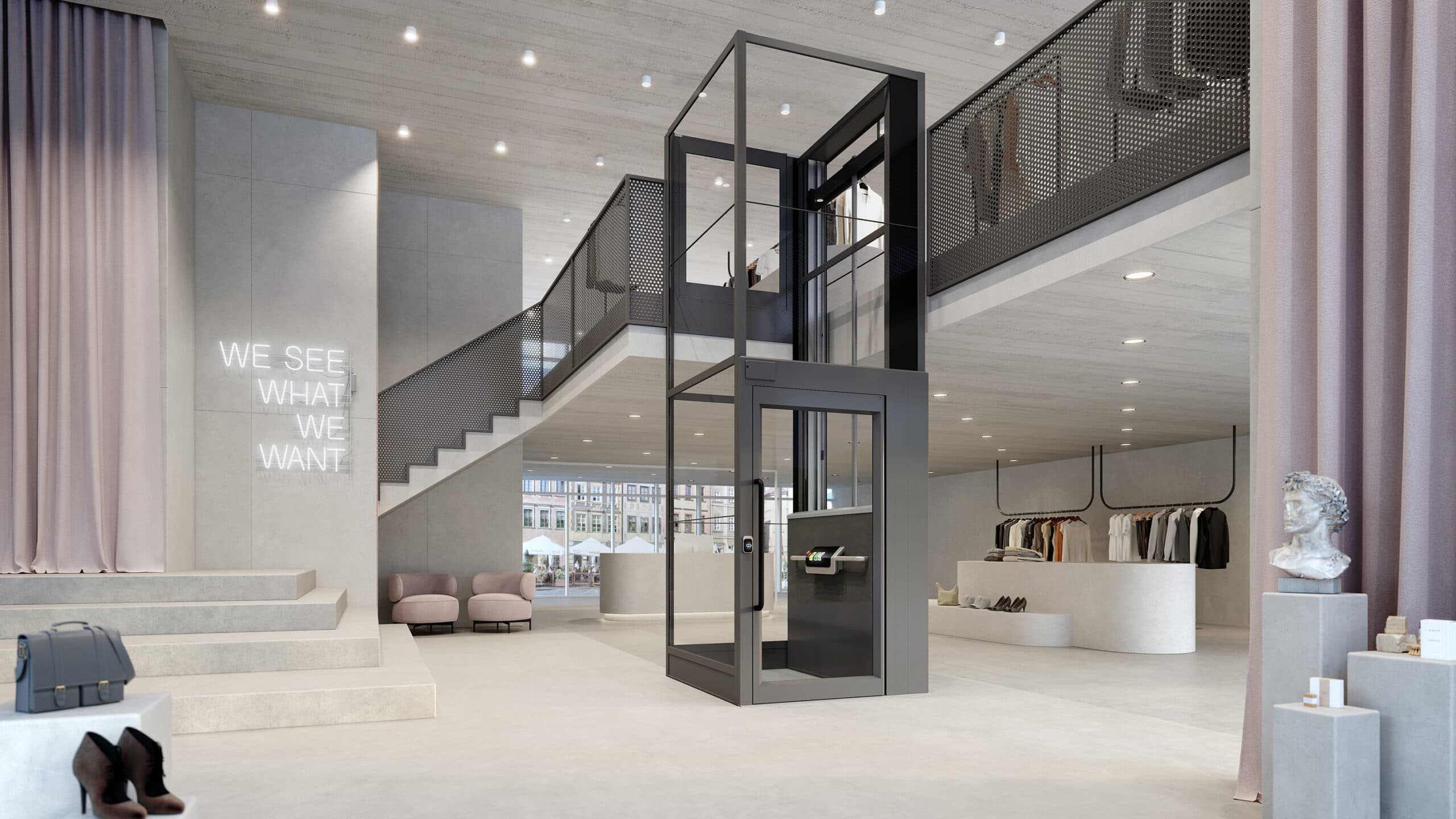 Grå Kalea Luna plattformshiss med glasschakt och grind på översta våningen, installerad mot ett entresolplan i en rymlig klädbutik med betongväggar, gammelrosa textildetaljer och möbler samt en vit neonskylt med texten "We see what we want"